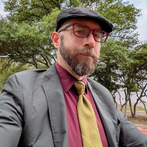 A photo of me, a 47 year old white man with a shaved head, a short cropped brown & grey beard, 

photographed from the chest up

In front of some live oak trees in an office park,

wearing

rectangular maroon acetate glasses, an olive green necktie in a trinity knot on a maroon dress shirt under a dark grey sport coat with a dark grey flat cap