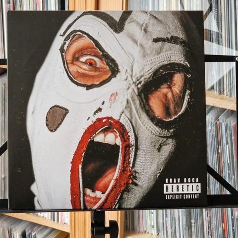 Das Bild zeigt ein Albumcover mit dem Titel "HERETIC" von Krav Boca. Es zeigt eine Nahaufnahme einer Person, die eine abgenutzte weiße Maske mit roten Akzenten und einem übertriebenen Ausdruck trägt, die weit geöffnete Augen und einen offenen Mund zeigt und rohe Emotionen betont.