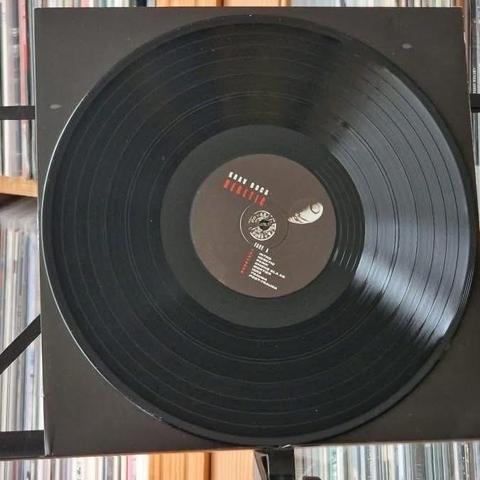 Das Bild zeigt eine Schallplatte, die auf einem Plattenspieler ruht, mit einem dunklen Etikett in der Mitte mit Text. Im Hintergrund sind Regale mit anderen Schallplatten sichtbar.