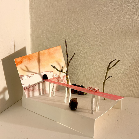 A small scale sculpture made of paper, with a bridge going toward a wall and through a tunnel, over trees and rocks. A car is on that pink road.