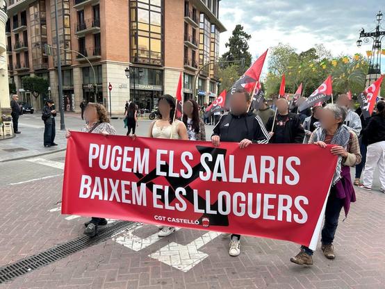 La imagen muestra una manifestación. Un grupo de personas (cuyas caras están difuminadas para proteger su identidad) está sosteniendo una pancarta grande y roja en medio de la calle. El texto en la pancarta está escrito en valenciano y dice: “PUGEM ELS SALARIS, BAIXEM ELS LLOGUERS”. Debajo del texto aparece el nombre del sindicato: CGT Castelló.

Detrás, más manifestantes marchan con banderas rojas y negras con las letras “CGT”.