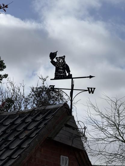 A decorative weather vane featuring Paddington bear - wearing a duffle coat and hat, positioned atop a roof. The vane includes directional indicators for north and west, set against a cloudy sky.