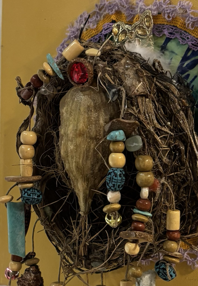 A goddess figurine made from a milkweed pod and cabochon sits inside a bird’s nest. Strands of beef’s hand from the nest. 