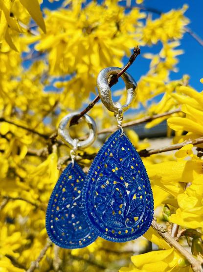 A pair of intricate blue teardrop earrings hangs from a branch against a backdrop of vibrant yellow flowers and a clear blue sky.