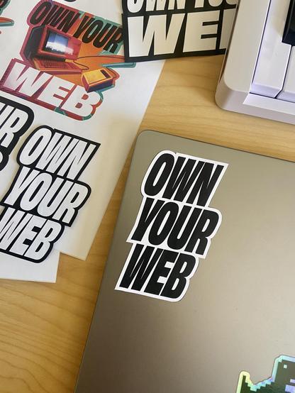 Paper versions of “Own Your Web” stickers next to a MacBook lid. One of them is placed on top of the computer.