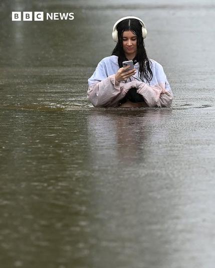 A girl with phone and headphones looking at her phone. 
Under the rain, half immersed in water.