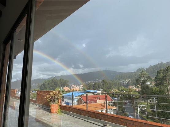 A view out my living room picture windows, out across the sunlit patio, looking southeast across the low, red brick buildings and trees below, towards the hills and cloudy grey sky.  A vivid rainbow curves down from beyond the frame the left, down to the buildings below, and a fainter rainbow arcs outside it.