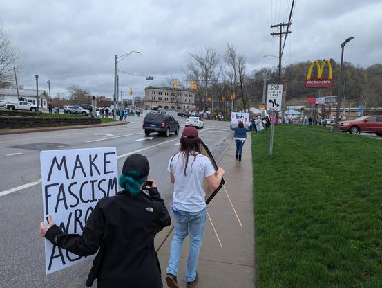 A small group of people walking towards a crowd of protesters while holding signs calling out the Trump administration.