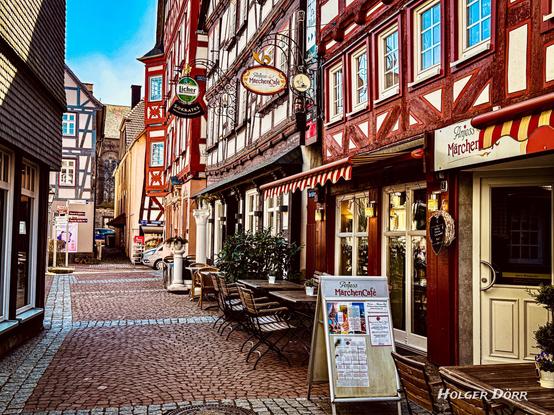 Romantische Fachwerkgasse in der Altstadt von Grünberg mit dem liebevoll dekorierten „Anjess MärchenCafé“. Vor dem Café stehen einladende Tische auf dem Kopfsteinpflaster, überragt von kunstvoll verzierten Fassaden und nostalgischen Wirtshausschildern. Im Hintergrund ragt eine alte Kirche empor – eine Szenerie wie aus einem Märchenbuch