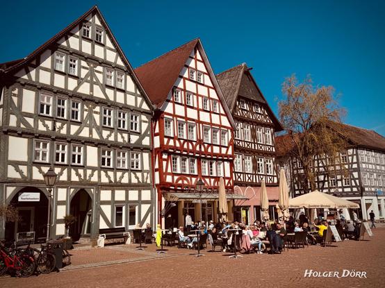 Historische Fachwerkhäuser säumen den sonnenbeschienenen Marktplatz von Grünberg. Menschen genießen das warme Frühlingswetter in einem Straßencafé, während der blaue Himmel und die lebendige Atmosphäre ein Gefühl von Leichtigkeit und Lebensfreude vermitteln.