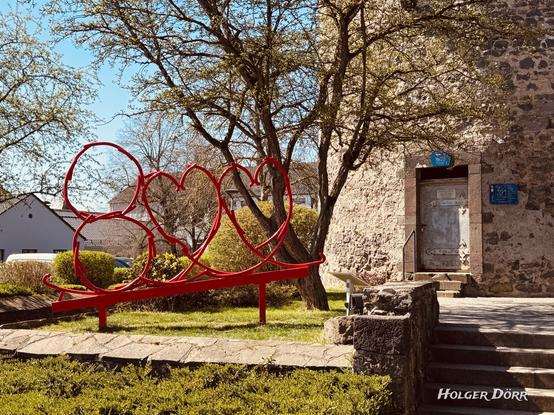 Moderne rote Skulptur in Form der Zahl “800” vor dem historischen Diebsturm in Grünberg. Die Skulptur wurde anlässlich der 800-Jahr-Feier der Stadt im Jahr 2022 errichtet und symbolisiert das Stadtjubiläum. Sie steht auf einer kleinen Grünfläche, umgeben von zart austreibenden Frühlingsbäumen. Das alte Gemäuer des Turms bildet einen kraftvollen Kontrast zur modernen Skulptur – ein stiller Dialog zwischen Geschichte und Gegenwart.