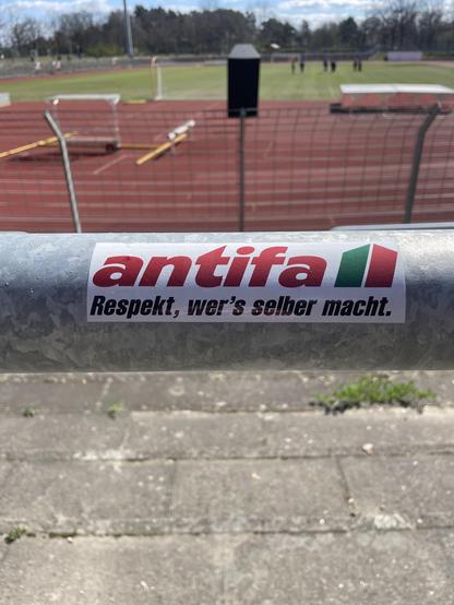 Aufkleber auf einem Geländer im Toom Baumarkt Stil: 
„antifa
Respekt, wer‘s selber macht.“
Im Hintergrund ist das Spielfeld im Mommsenstadion zu erkennen.