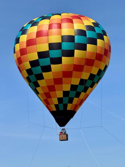 A colorful hot air balloon with a checkered pattern is flying against a clear blue sky, featuring a basket below with several people inside.