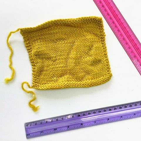 A slightly wrinkled yellow knitted swatch in stockinette stitch is shown on a white background. Two rulers are also visible.