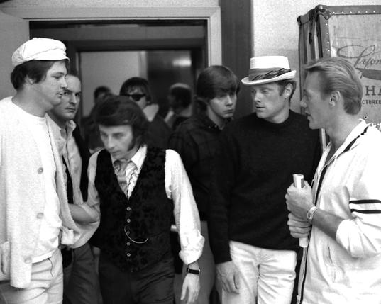 Pic from the mid-60s showing Brian Wison, Mike Love and Al Jardine of the Beach Boys casting sidelong looks at Phil Spector, who touches Brian's arm as he walks by him. There are several other presumably music-biz types in the room.