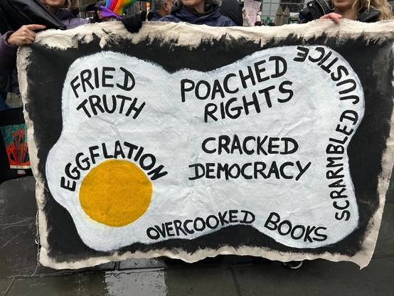 Image of a blanket at a protest in NY with the phrases "fried truth", "poached rights", "eggflation", "cracked democracy", "overcooked books", and "scrambled justice" on it.