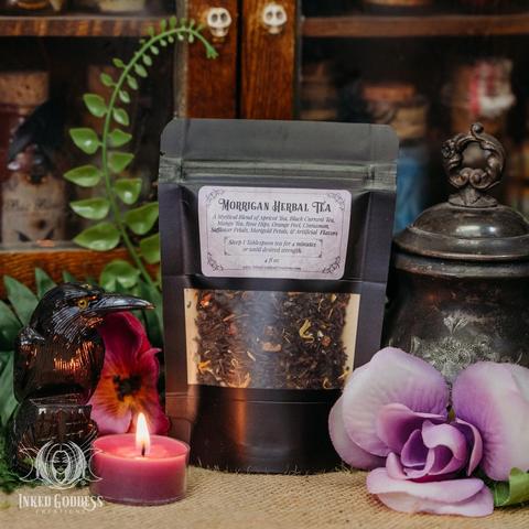 A black pouch of dried herbs sits on a burlap-covered surface. There is a window in the front and a label that reads, "Morrigan Herbal Tea" with a list of ingredients and instructions. The imagery includes. This is an exclusive product from Inked Goddess Creations.