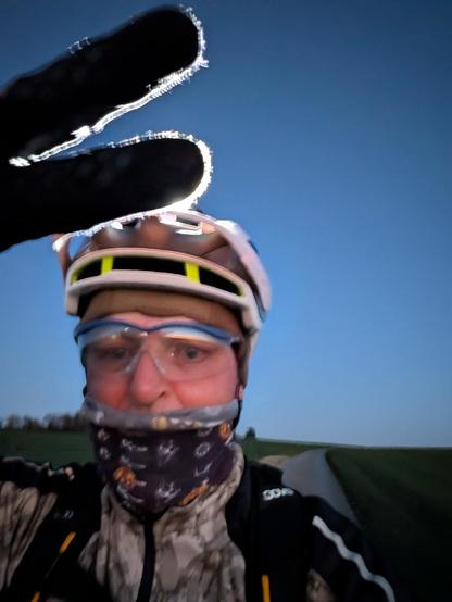 Das Bild zeigt eine Person, die auf einem Fahrrad unterwegs ist. Die Person trägt einen weißen Radhelm mit gelben Akzenten und eine schwarze Handschuhe, die an den Fingerspitzen mit Eiskrystallen bedeckt sind, was auf kalte Temperaturen hinweist. Sie hat eine Gesichtsmaske mit einem Muster aus verschiedenen Symbolen und ein Paar transparente Sicherheitsbrillen. Die Person trägt eine Kombination aus einem dunkelgrauen und beigefarbenen Jacken mit einem schwarzen Rucksack. Im Hintergrund ist ein offenes Feld mit einer asphaltierten Straße zu sehen, die sich in die Ferne erstreckt. Der Himmel ist klar und blau, was auf einen klaren Tag hinweist.
