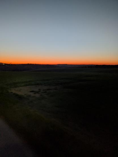 Das Bild zeigt eine Landschaft bei Sonnenuntergang. Der Himmel ist in einem warmen Orange und Rot gefärbt, während die oberen Bereiche in einem kühleren Blau gehalten sind. Im Vordergrund erstreckt sich ein grasbewachsener Feldzug, der sich bis zum Horizont erstreckt. Die Landschaft ist flach und weitet sich aus, mit sanften Hügeln im Hintergrund. Am Horizont sind einige Lichter sichtbar, die auf eine Entfernte Siedlung hinweisen. Die Szene ist ruhig und friedlich, mit einem Gefühl der Abgeschiedenheit.