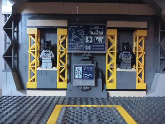 The rear wall of the Batcave, with Batsuit gallery and Gotham City computer screens, and a vehicle plate on the floor