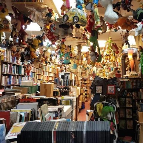 A photo inside Bonnett's Bookstore.
A row of DVDs in the foreground partially obscures a Marvin the Martian shoe caddy hanging from an old-school hand-crank pencil sharpener. To the right is a dimly lit alcove of bookshelves, above which can be seen an illuminated Leg Lamp. To the left of Marvin is a bright aisle full of shelves and boxes of books stretching into the distance. Further left, it's evident there's another aisle extending beyond a set of floor-to-ceiling bookshelves, hinting at further stacks to explore. Throughout the shot are thousands of toys decorating shelves and hanging above aisles.