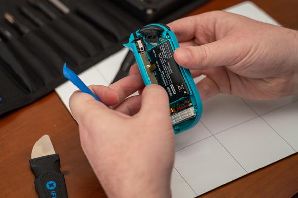 Two hands separating the outer shell from a blue Nintendo Switch Joy-Con. They're holding it partially open above a white magnetic project mat on a wooden table. On the table is also an iFixit Jimmy and Pro Tech toolkit in the background, with the screwdriver from that kit sitting on the project mat. The left hand's index finger is also gripping an iFixit Opening Tool.