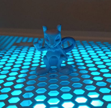 Foto de uma miniatura do pokemon Mewtwo em cima de uma estrutura de metal com diversos furos em formato circular, de onde sai uma luz azul claro, que o ilumina.