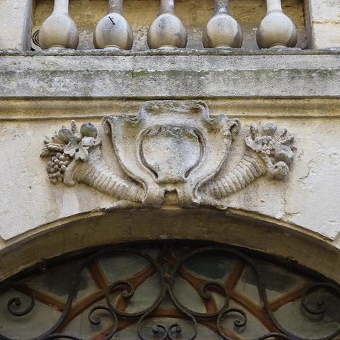 façade en pierre avec un blason sculpté et une corne d'abondance de chaque côté. Au dessus on aperçoit la partie basse d'un balcon en pierre, en dessous le haut d'une porte arrondie avec des volutes en fer forgé.