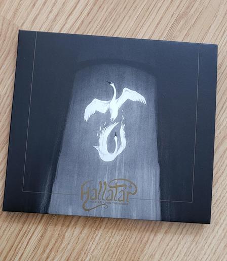 A CD album on orange-brown flooring. Album cover is mostly black, but a broad strip of light gray is painted in the middle, it looks like a beam of light, a window or drapes. Inside the strip there are two white swans, the one above is flying, but the one under the first is falling down and its wings, swept upward, are losing feathers. There is a thin dark gold border about 13mm from all edges except the upper one. Band name Hallatar is in the middle of the golden border at the lower edge in swirly, beautiful dark gold letters and a star within the swirl that starts from the R and ends up crossing the T. Album name "No stars upon the bridge" can be barely made out in tiny gold print letters under band name.