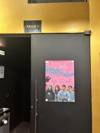 Sur la photographie nous pouvoir une porte noire à demi ouverte avec un mur très jaune. Sur une moitié de cette porte est collée une affiche au fond rose dont le titre bleu apparaît : GIRLS FOR TOMORROW. Nous pouvons voir quatre filles une fille noire à gauche une fille aux veux ondulés à sa droite, puis une filles les bras croisés et les cheveux attachés et enfin une fille au cheveux courts. On peut également voir des écritures, des signes noirs à peu près partout sur l'affiche et  Donal Trump à gauche en train de faire un doigt d'honneur mais il est barré par une sorte d'encre de stylo.