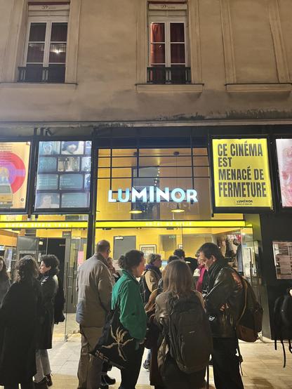 Sur la photographie, nous pouvons voir l'extérieur d'un cinéma avec l'inscription Luminor allumée d'une lumière blanche. Il semble faire plutôt sombre signalant une fin de soirée. Nous voyons à l'intérieur du cinéma, des murs très jaunes.  Plusieurs groupes de personnes se trouvent devant le cinéma certaines et certains portant des sacs et sac à dos. Le signe "Luminor" est au milieu d'autre affiches dont une seule qui est visible sur la photo. Cette affiche a un fond jaune et des écritures noires. Il y est écrit "ce cinéma est menacé de fermeture" 