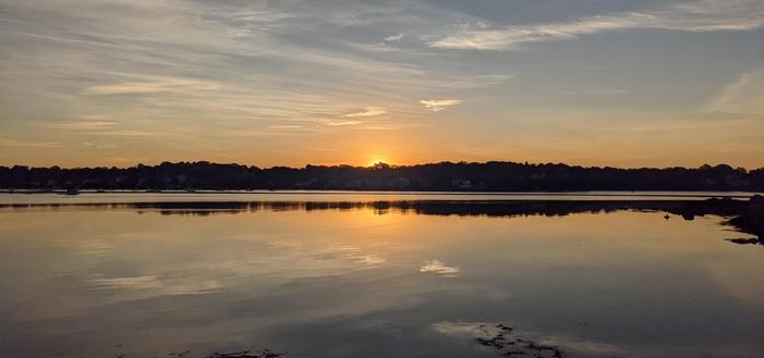 Historical Documentation in the form of a photograph of a rising sun over the ocean bay at the Historic Pioneer Village in Forest River Park, Salem, MA. The rising sun warms up the sky as it creeps over the treeline on the horizon.  It's warm glow is mirrored in the water.  There are just a few wisps of clouds in the blue sky.  It's the dawn of a brand new day where anything could be and is possible.  