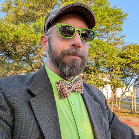 A photo of me, a 47 year old white man with a shaved head, a short cropped brown & grey beard, 

photographed from the chest up

In front of some live oak trees in an office park,

wearing

rectangular lime green acetate sunglasses, an golden brown textured bow tie on a lime green dress shirt under a dark olive green sport coat with a tan flat cap