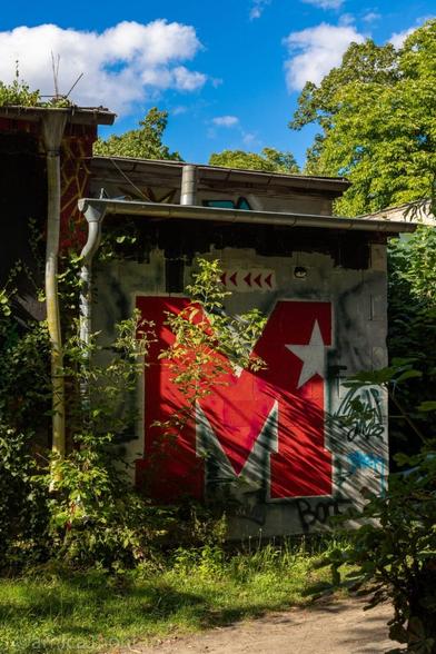 A red letter "M" with a white star is prominently displayed on a wall covered in graffiti, surrounded by greenery and under a bright blue sky with scattered clouds.