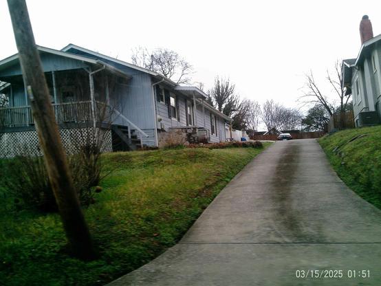 A driveway leading up between a light blue house and a gray house, with grassy areas and bare trees.