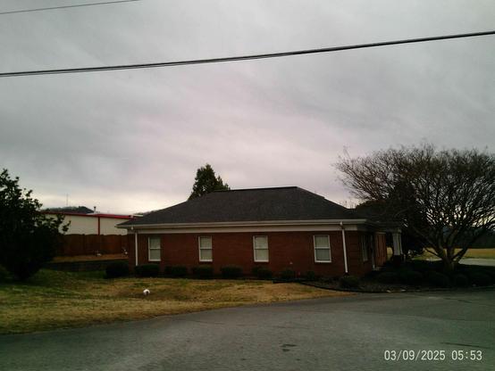 Brick building with dark gray roof and several windows, situated on a paved lot next to a fenced yard.