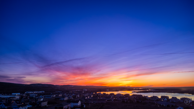 Aerial view of Schefferville at sunset, captured on June 7, 2022. The sky displays a breathtaking gradient of deep blues, purples, and fiery oranges as the sun sets behind the distant hills. Below, the town's houses and buildings are nestled along a calm river, reflecting the golden hues of the fading daylight. Wisps of clouds stretch across the sky, enhancing the tranquil atmosphere as night falls over Schefferville.