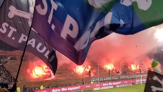 Bildbeschreibung von KI:
Das Foto zeigt eine emotionale und energiegeladene Szene aus dem Millerntor-Stadion, der Heimstätte des Fußballvereins FC St. Pauli in Hamburg.

Im Vordergrund sind große Fahnen mit dem Schriftzug „ST. PAULI“ und dem Totenkopf-Logo zu sehen – ein bekanntes Symbol der Fanszene. Im Hintergrund lodern bengalische Feuer (Pyrotechnik), was die Stimmung zusätzlich anheizt. Über der Tribüne ist ein riesiges Banner gespannt.

Auf dem Zaun vor der Kurve ist ein Schriftzug zu lesen:
„VORAN FC SANKT PAULI – NIEMAND ZWINGT DICH IN DIE KNIE“ – ein typischer ironischer bis kämpferischer Fangesang.

Auch sichtbar sind Werbebanden (z. B. Astra. Was dagegen?) und einige Ordner in gelben Westen. Die Szene ist offensichtlich aus dem Fanblock aufgenommen worden, inmitten der Choreografie, vermutlich kurz vor oder während des Spiels.
