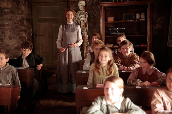 Auch dieses Bild stammt aus dem Film „Louise und die Schule der Freiheit“ und zeigt eine typische Unterrichtsszene in der Dorfschule:

Im Mittelpunkt steht erneut Louise, die Lehrerin, die freundlich lächelt und mit einem Zeigestock in der Hand hinten im Klassenraum steht. Hinter ihr ist ein Skelett-Modell zu sehen – ein klassisches Unterrichtsobjekt für den Biologieunterricht.  

Vor ihr sitzen mehrere Kinder an alten Holzpulten, die teils aufmerksam, teils fröhlich in die Kamera oder zu ihr schauen. Besonders auffällig ist das warme, weiche Licht, das die Szene nostalgisch wirken lässt – wie eine Erinnerung an vergangene Zeiten.

Der Raum selbst wirkt rustikal und einfach: grobe Steinwände, eine hölzerne Tür, ein offenes Regal mit alten Büchern und Utensilien – das Setting unterstreicht die ländliche und einfache Atmosphäre.  

Die Szene vermittelt Wärme, Gemeinschaft und den Geist einer alternativen, kindgerechten Pädagogik – ganz im Sinne des Filmtitels *„Schule der Freiheit“*.