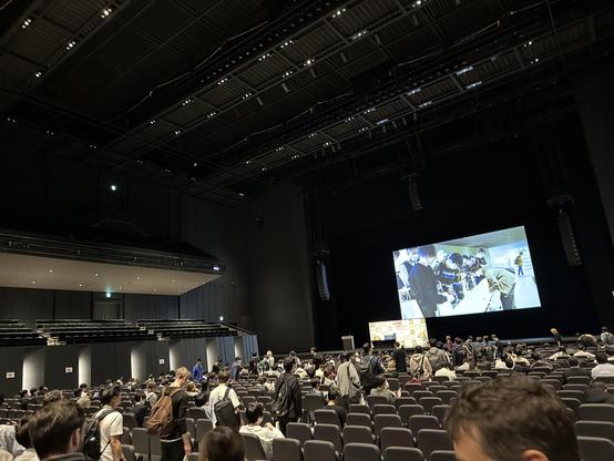 Black venue with screen at the front. People are lining up in the aisles to find a seat.