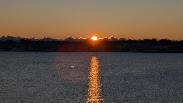 Historical Documentation in the form of a photograph of a rising sun over the ocean bay in Salem, MA. The sun is rising in the background over some Cumulus Clouds resting between the sun and the trees on the horizon. The sun is peeking over their fluffy peaks casting a warm orange glow over the horizon. The water is calm and still, reflecting the colors of the sky. In the foreground, there is a small island with trees and buildings visible in the distance. The sky is a mix of orange and yellow hues, with the sun partially obscured by the clouds. The overall mood of the image is peaceful and serene as ducks swim silently in the calm waters.