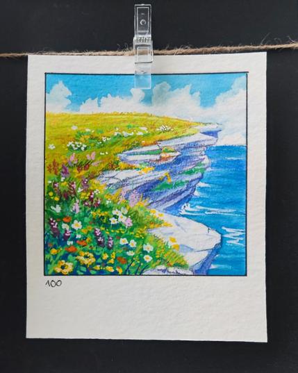 Square watercolor framed with a black line on a rectangular, portrait-oriented paper. A coastal cliffside with layered, weathered rocks jutting out over the ocean. The cliff is covered in lush green grass and a dense spread of colorful wildflowers in shades of yellow, white, purple, and pink. The deep blue ocean stretches to the horizon, with gentle waves lapping against the rocks below. Above, the sky is a vibrant blue, dotted with fluffy white clouds. Number 100 is written in the bottom left corner.