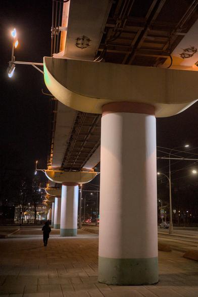 The monorail goes into the distance. View from below. Sturdy supports. Late evening. Lights are burning. A silhouette of a man can be seen next to one of the supports.