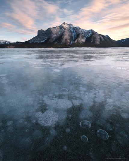 A mountain sits in the evening sun as sunset starts. High stratospheric clouds are lit softly pink. The frozen lake in the foreground is full of white and opague filled bubbles creating amazing little patterns in the ice.
