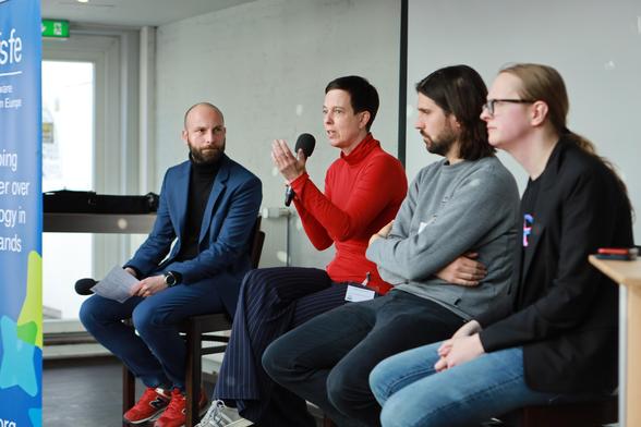A woman on a panel with three other persons holding a microphone and talking to an audience.