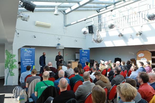 Image from the back of a room full of people with two people standing and presenting between two roll ups.