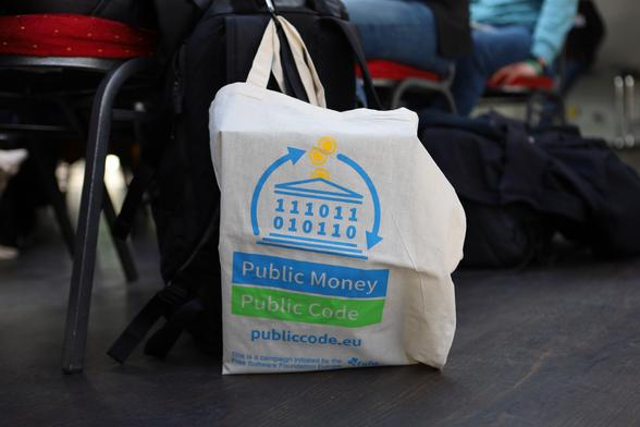 A tote bag reading Public Money? Public Code! and the URL publiccode.eu leaning on a audience chair
