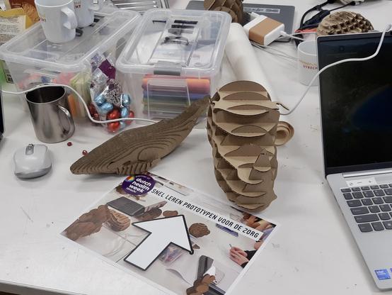 Tafel met kartonnen modellen, laptop, muis, dozen met stiften, en een A4 met titel: SNEL LEREN PROTOTYPEN VOOR DE ZORG