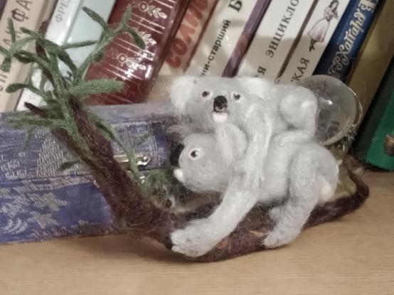 There is a craft on the bookshelf. This is miniature interior toy, made from nettle fibers by dry felting, in the form of a koala with a baby. An adult koala sits on a eucalyptus branch and holds a branch with one paw and eats leaves. The baby clings to the mother koala's back with all paws, its head is turned to the side. Thank you for your attention!