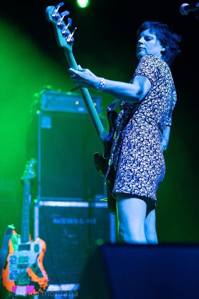 A musician plays the bass guitar on stage, wearing a floral dress. The background features green lighting and amplifiers. An electric guitar in bright colors is positioned nearby.